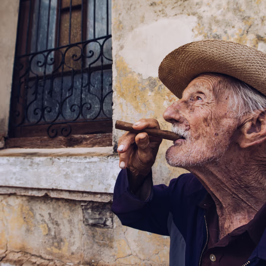 Пожилой кубинец наслаждается сигарой сидя у стены старого здания