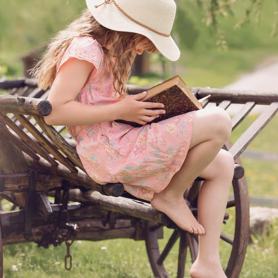 Маленькая девочка читает книгу сидя на деревянной телеге в саду