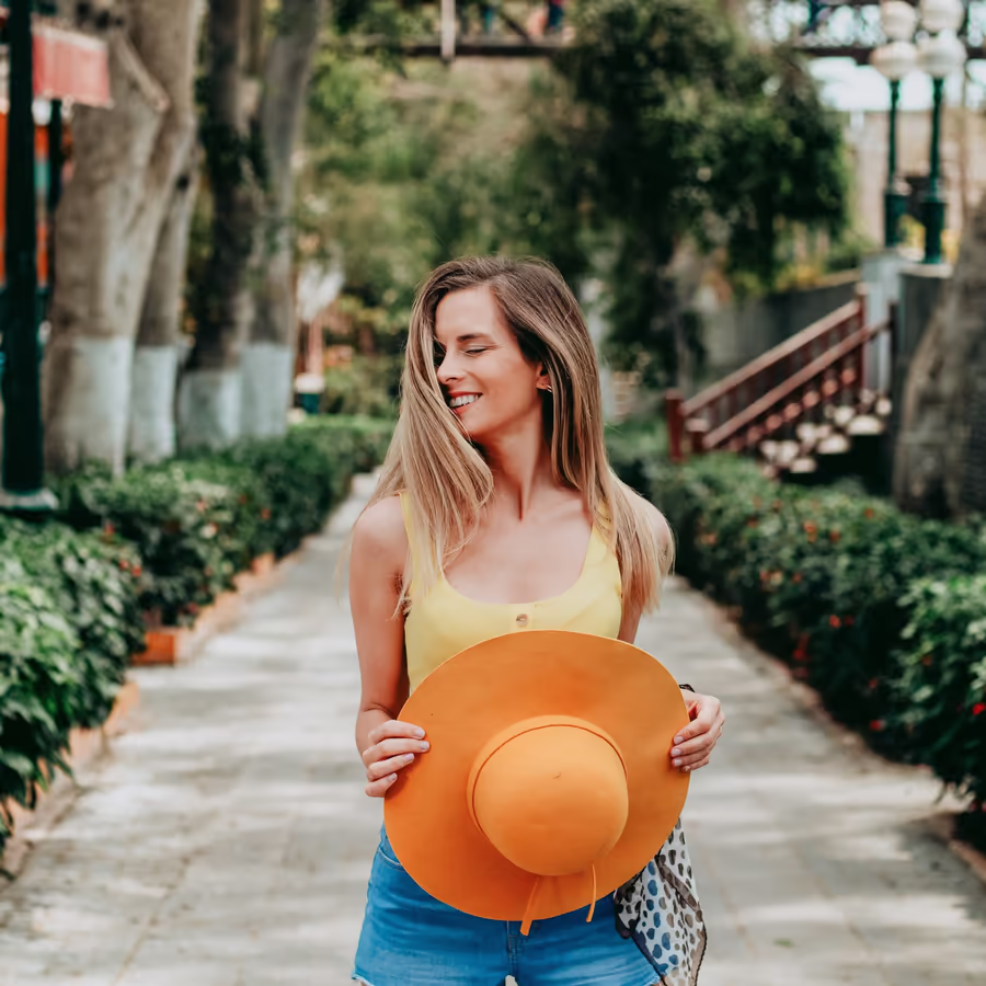 Молодая женщина держит большую оранжевую шляпу улыбаясь на фоне зелени